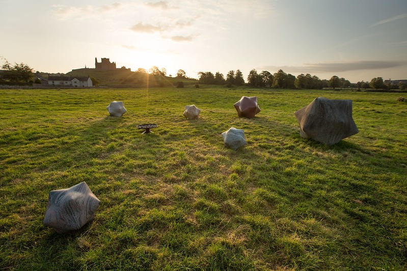 An outdoor installation of star shaped rock sculptures of various sizes scattered across a green field at sunset